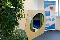 Photographie d'un cocon pour pratiquer la micro-sieste : cocon en bois, aménagé pour pouvoir se reposer. A gauche du cocon, il y a une plante verte et à droite, un kakémono explicatif.