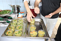 Photographie d'un cours de cuisine : deux personnes déposent des récipients garnis dans un contenu en métal.